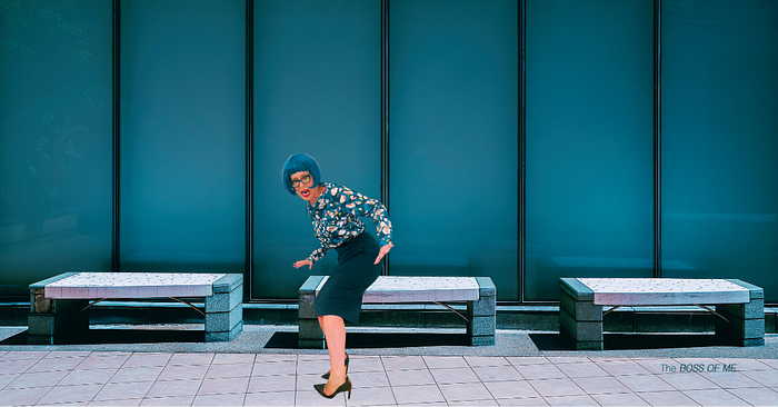 A woman with blue hair mid-stumble in high heels on a sidewalk near an office building.