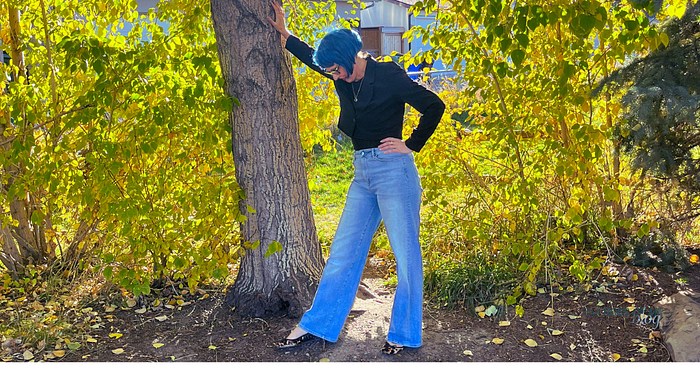 A woman with blue hair wearing leopard print flats, a black top, and a black blazer, posing next to a tree outdoors.
