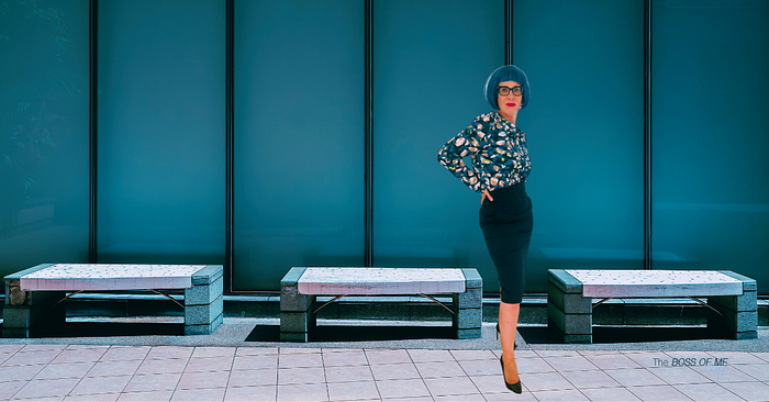 A woman with blue hair wearing a patterned blouse, a pencil skirt, and black high heels, standing in front of an office building.