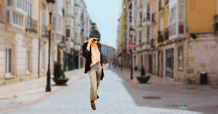 A woman with blue hair dressed in leopard print boots, jeans, a white T-shirt, and a black blazer, standing outdoors.